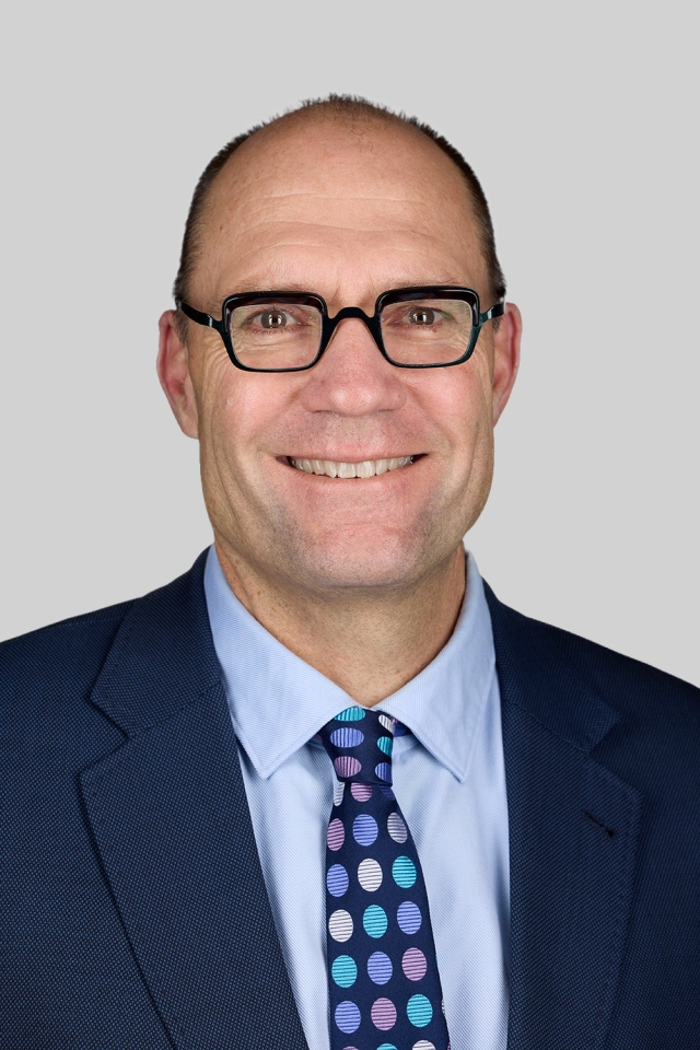 Studio‑style head‑and‑shoulders portrait of a person wearing rectangular glasses, a blue suit jacket, light blue collared shirt, and a patterned tie, photographed against a plain gray background.
