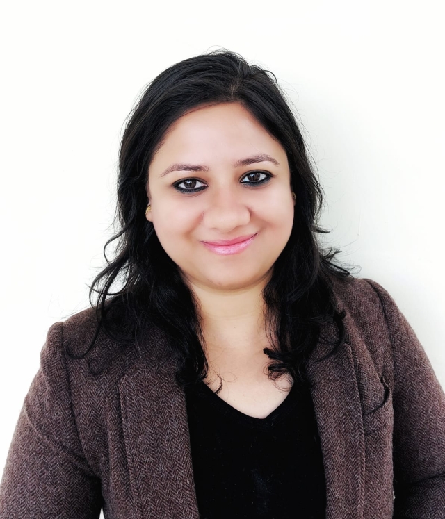 Studio‑style head‑and‑shoulders portrait of a person with shoulder‑length dark hair, wearing a brown blazer over a dark top, photographed against a plain white background.