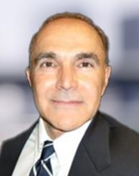 Head‑and‑shoulders portrait of a person in a dark business suit with a white collared shirt and patterned tie, photographed against a softly blurred background.