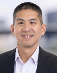 Professional head‑and‑shoulders portrait of a person wearing a dark suit jacket over a light collared shirt, photographed against a softly blurred indoor background.