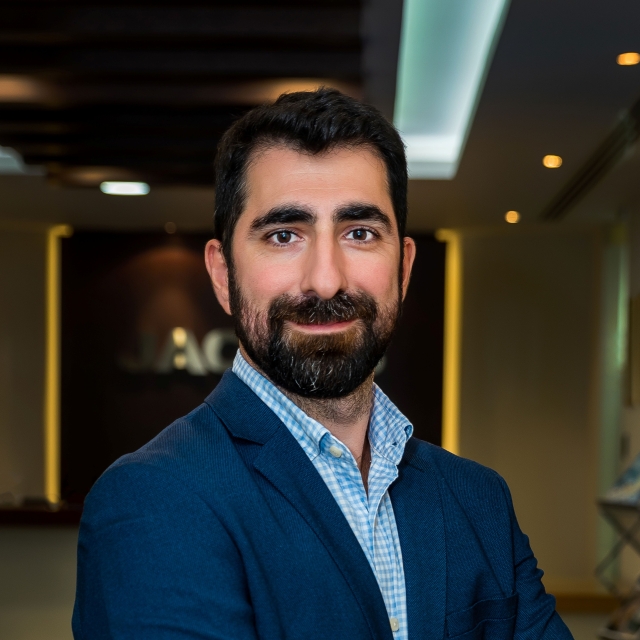 Head‑and‑shoulders portrait of a person with short dark hair and a beard, wearing a dark blazer over a light blue checkered shirt, photographed indoors with a softly blurred background.