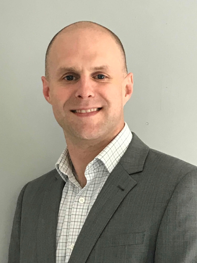 Studio‑style head‑and‑shoulders portrait of a person wearing a gray suit jacket over a light checkered collared shirt, photographed against a plain light gray background.