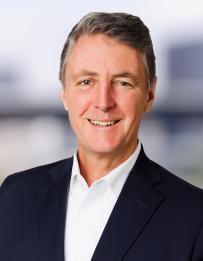 Head‑and‑shoulders portrait of a person in a dark business jacket and white collared shirt, photographed against a softly blurred outdoor or architectural background.