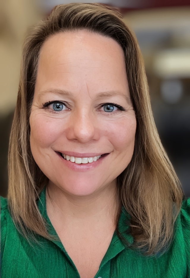 Studio‑style head‑and‑shoulders portrait of a person with shoulder‑length hair wearing a green blouse, photographed against a softly blurred interior background.