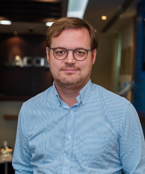 Portrait of a person wearing round glasses and a light blue striped shirt, standing inside a modern office with a softly blurred background.