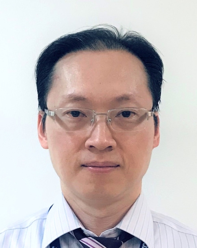 Studio‑style head‑and‑shoulders portrait of a person wearing rimless glasses, a light striped collared shirt, and a dark tie, photographed against a plain white background.