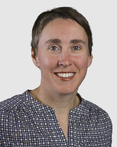 Head‑and‑shoulders studio portrait of a person in a patterned, light‑colored blouse, photographed against a plain light gray background.