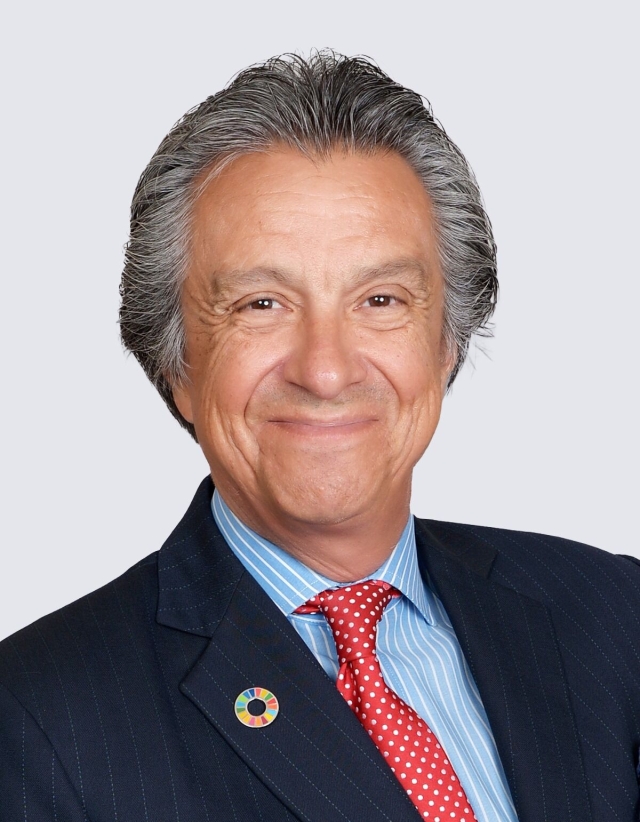 Head‑and‑shoulders studio portrait of a person in a dark business suit with a patterned tie and collared shirt, photographed against a plain light background.