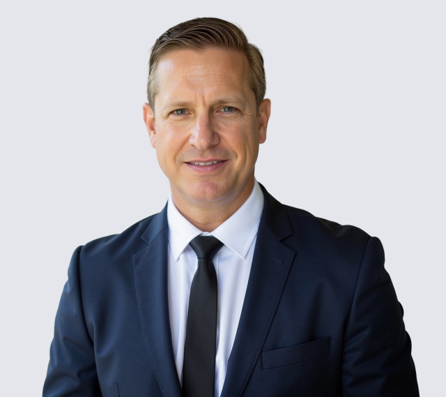Head‑and‑shoulders portrait of a person in a dark business suit with a white collared shirt and black tie, photographed against a plain light gray background.
