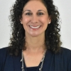 Headshot of a smiling woman with dark brown, curly hair wearing a navy blue shirt and silver necklace