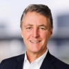 Head‑and‑shoulders portrait of a person in a dark business jacket and white collared shirt, photographed against a softly blurred outdoor or architectural background.