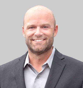 Head‑and‑shoulders studio portrait of a person wearing a dark blazer over a light‑colored collared shirt, set against a plain gray background.