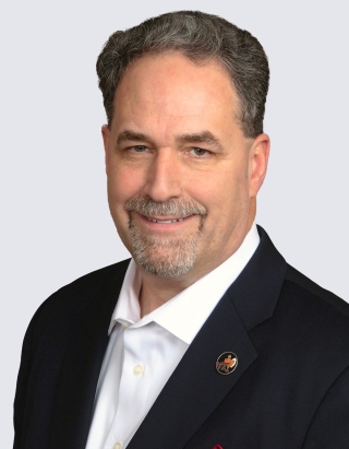 Head‑and‑shoulders studio portrait of a person in a dark business jacket with a white collared shirt, photographed against a plain light gray background.